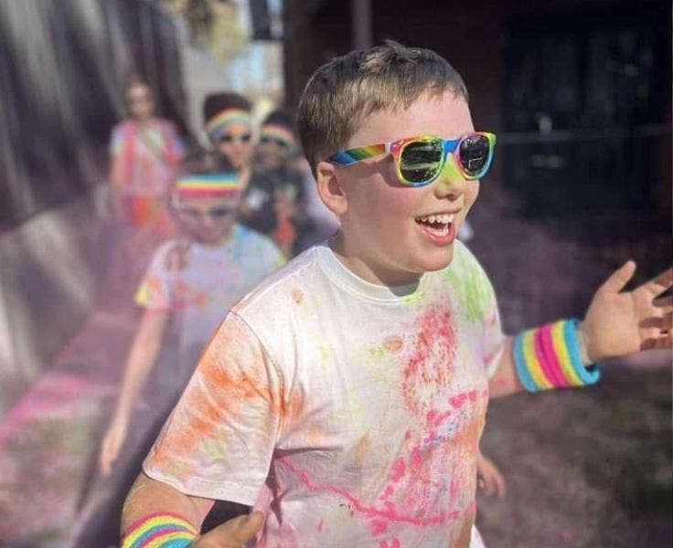 Photo of a very happy student covered in coloured powder and wearing rainbow glasses, running toward the camera.