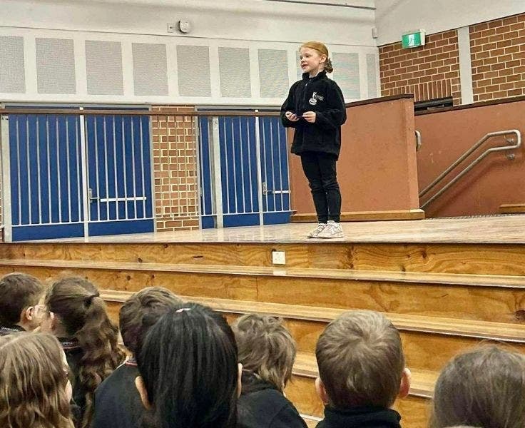 Photo of a young student on a stage in a public speaking competition with an audience below listening.